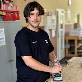Junger Mann mit braunen Haaren schleift ein Stück Holz.