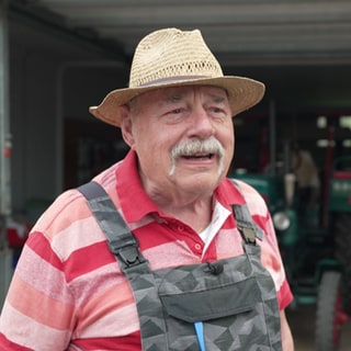 Dietmar Beck hat einen Strohut auf und eine Latzhose an. Er steht vor seiner Garage, die voll mit Traktoren ist.