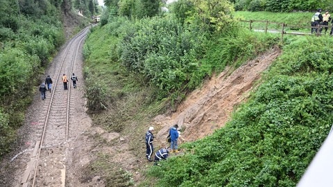 Erde liegt nach einem Hangrutsch auf den Gleisen, im Hintergrund ist der entgleiste Regionalexpress auf der Strecke zu sehen. Bei dem Zugunglück im Südosten Baden-Württembergs sind drei Menschen ums Leben gekommen. 