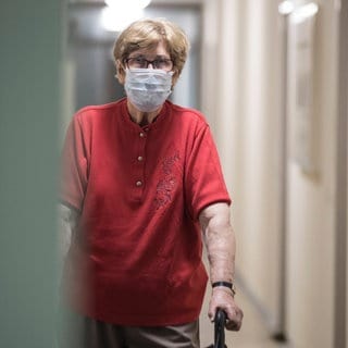 Eine Seniorin mit OP-Maske und Rollator geht alleine über einen Flur im Altenheim: Altenheime wollen und sollen ihre Bewohner vor einer Corona-Infektion bewahren. Doch dieser Schutz hat Folgen für alle, die teils monatelang allein in ihren Zimmern lebten: Einsamkeit, Depressionen, ein schwindender Lebenswille. 