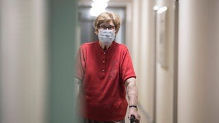 Eine Seniorin mit OP-Maske und Rollator geht alleine über einen Flur im Altenheim: Altenheime wollen und sollen ihre Bewohner vor einer Corona-Infektion bewahren. Doch dieser Schutz hat Folgen für alle, die teils monatelang allein in ihren Zimmern lebten: Einsamkeit, Depressionen, ein schwindender Lebenswille. 