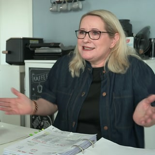 Mechaela, eine 57-jährige, blonde Frau, sitzt an einem Tisch. Vor ihr liegt ein Ordner mit Abrechnungen. Sie hebt ihre Arme und signalisiert Hilflosigkeit.