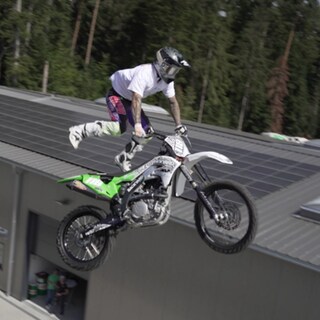 Freestyle-Motocrosser André Mikitta fliegt in waghalsiger Pose mit seinem Motorrad durch die Luft, vor blauem Himmel und unter Applaus des Publikums.