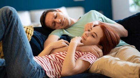 Junges Paar kuschelt auf dem Sofa: Bindungshormon, Treue- oder Orgasmushormon: Oxytocin werden Superkräfte nachgesagt. Zur Superkraft für unser Wohlbefinden wird es aber nur, wenn wir mit einem anderen Menschen zugewandt in Beziehung stehen. 