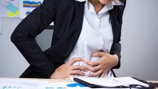 Frau in Business-Outfit sitzt am Schreibtisch und hält sich mit schmerzverzerrtem Gesicht den Bauch. Was sind Ursachen für starke Blähungen? Welche Hausmittel helfen?