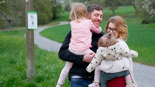 Jenny, ihr Ehemann Sascha und ihre beiden Töchter stehen im Feld. Hinter ihnen ist ein Weg, Bäume und Gras. Sascha hält Ella und Jenny hält Lea im Arm.