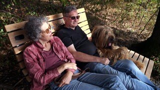 Inga und Claus Berg sitzen mit ihrem Hund auf einer Bank im Wald.