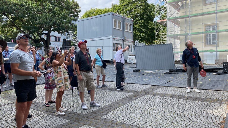 Touristen stehen auf der Baustelle auf dem Florinsmarkt mitten in der Koblenzer Altstadt und warten darauf, dass der Augenroller seine rote Zunge heraussteckt.