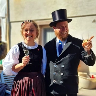 Bettina und Gabriel Wagner stehen vor ihrem Haus. Sie tragen Tracht und halten Sektgläser in der Hand.