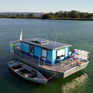 Hausboot „Netta“ schwimmt mitten im Bodensee zwischen Wasservögeln.