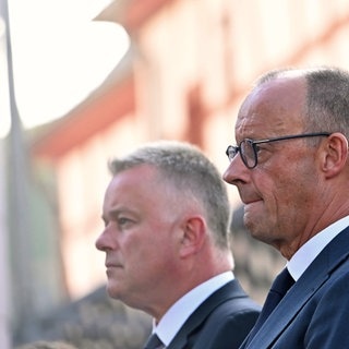Bundeskanzler Friedrich Merz (CDU) beißt die Lippen aufeinander - im Hintergrund steht der rheinland-pfälzische Ministerpräsident Gordon Schnieder (CDU).