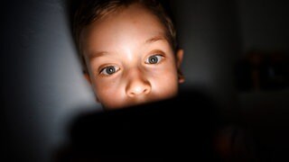 Symbolfoto: Gestellte Aufnahme zum Thema Mediennutzung bei Kindern. Ein Kind schaut im Dunkeln auf das Display eines Smartphones.