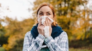 Eine Frau steht vor einem Baum und putzt sich die Nase. Die Pollen-Saison beginnt immer früher.