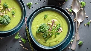 Grüne Brokkoli-Suppe in blauem Schälchen auf grauer Beton-Platte. Wird Brokkoli roh genossen, ist er besonders gesund. 