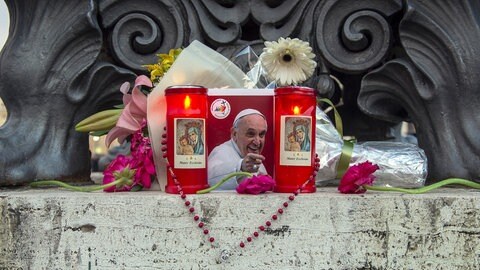 Ein Bild des verstorbenene Papstes Franziskus steht inmitten von Kerzen und Blumen. Ein Rosenkranz ist um das Bild geschlungen. Trauerfeierlichkeiten, Beisetzung, Papst-Wahl im Konklave – ein minutiös durchgeplantes Räderwerk ist nach dem Tod von Papst Franziskus im Vatikan in Bewegung. Vatikan-Experte Andreas Englisch erklärt es in SWR1 Leute im Detail.