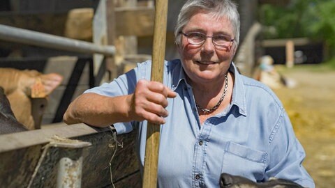 Frau mit blauer Jeansbluse steht an einem Stall und schaut in die Kamera.