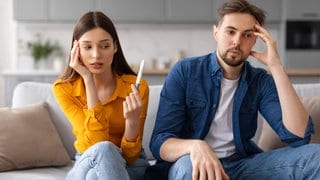 Eine junge Frau sitzt auf dem Sofa und hält mit nachdenklichem Blick einen Schwangerschaftstest in der Hand, während der Mann neben ihr besorgt und nachdenklich wirkt.