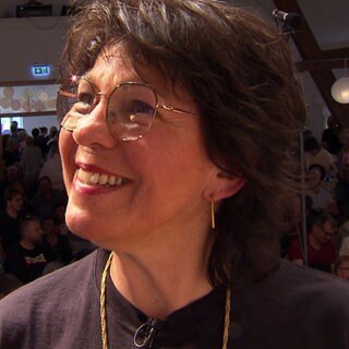 Ulrike Keßler, Leiterin des Kinderchor Bolstern, schaut schräg nach links oben und lächelt. Im Hintergrund sind im Dunkeln ein paar Menschen zu sehen.