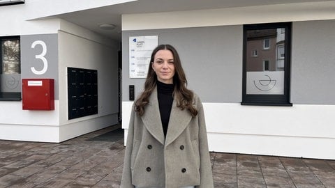 Eine junge Frau steht vor einer Hausarztpraxis. Sie hat über ein Jahr lang nach einem Rheumatologen gesucht.