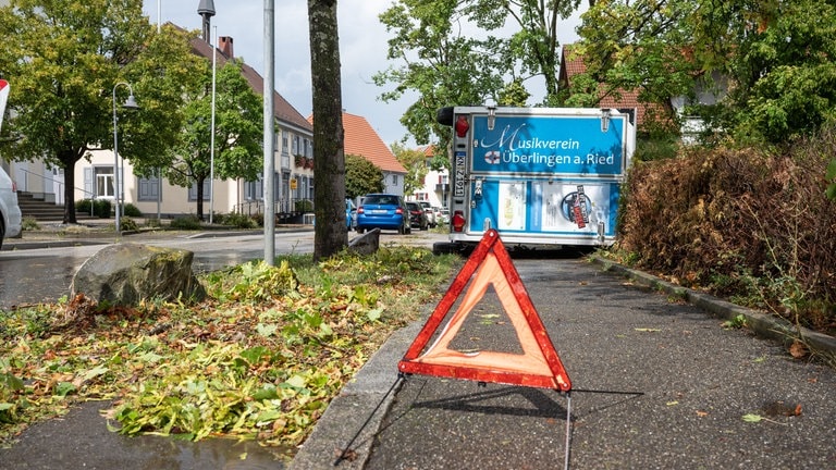 Ein Warndreieck steht vor einem umgestürzten Anhänger auf einem Gehweg in Überlingen am Ried (Kreis Konstanz) nach einem schweren Unwetter.