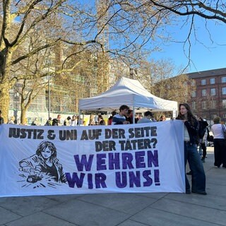 Zwei Demonstrierende halten ein Banner "Justiz & Uni auf der Seite der Täter? Wehren wir uns!"
