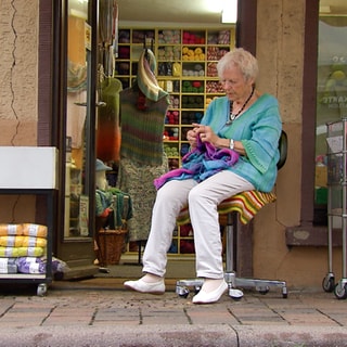 95 Jahre alte Frau sitzt vor dem Eingang ihres Wollgeschäfts auf einem Drehstuhl und strickt. Sie trägt einen gestrickten, blau-grünen Pullover und eine weiße Hose und weiße Ballerinas. Sie schaut auf das lila Strickzeug auf ihrem Schoß.