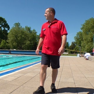 Alessandro Bagnoli trägt ein rotes T-Shirt und eine kurze schwarze Badehose. Er läuft auf der rechten Seite am Rand eines blauen Schwimmbeckens entlang.