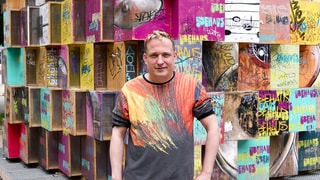 Jean-Marc ist Musiker. Er hat Tourette und räumt mit Vorurteilen auf. Er steht vor einer Wand, die aus bunten Würfeln besteht. Er schaut in die Kamera. 
