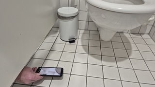 Ein Mann schiebt ein Handy unter der Trennwand einer Toilettenkabine hindurch.