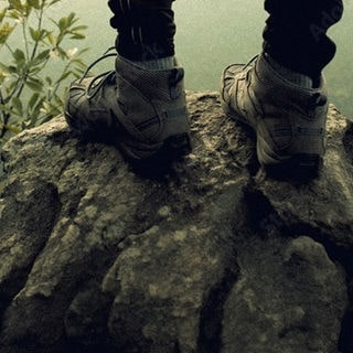 Bildausschnitt von Schuhen eines Wanderers, der auf einem schroffen Berggipfel steht, umgeben von nebliger, karger Landschaft.
