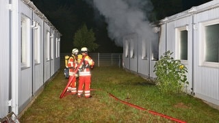 Zwei Feuerwehrmänner stehen vor einem Wohncontainer der Leonberger Sammelunterkunft. Aus den Fenstern raucht es. Eine Person ist bei dem Brand im Container ums Leben gekommen.