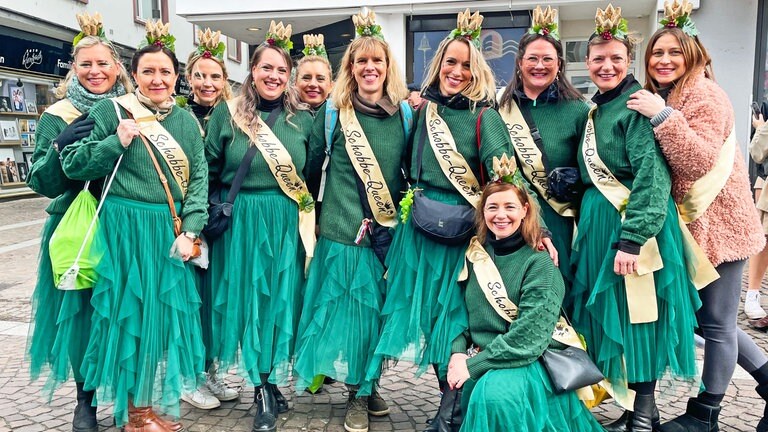 Eine Grupp junger Frauen trägt grüne lange Tüllröcke und grüne Oberteile. Alle tragen eine goldene Scherpe auf der steht: Schobbe-Queen. 