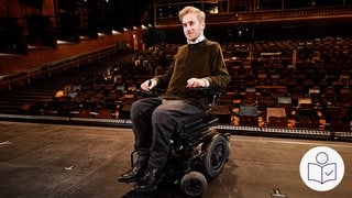 Der Schauspieler Samuel Koch auf einer Theater·bühne. Im Hintergrund ein leerer Zuschauer·raum. Rechts das SWR Logo für Leichte Sprache. 