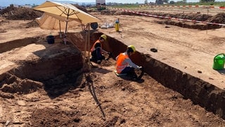 Zwei Arbeiter legen in einer Baugrube Erdschichten zur archäologischen Untersuchung frei