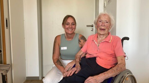 Eine junge Frau und eine alte Frau im Rollstuhl lächeln in die Kamera. 