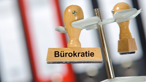 Ein Holzstempel ist beschriftet mit dem Aufdruck "Bürokratie"; dahinter ein Aktenschrank: Viele haben den Eindruck, dass die Verwaltung in Deutschland immer schlechter funktioniert. Frust und Ärger über Behörden nehmen zu. Dabei sind sie unverzichtbar – für jede und jeden Einzelnen und die Gesellschaft insgesamt.