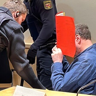 Ein Mann, der sein Frau getötet hat, hält sich im Gericht eine rote Mappe vor sein Gesicht.