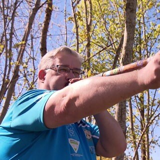 Thorsten Bouillon trägt ein blaues Hemd mit diversen Logos und hält ein Blasrohr aus Holz vor seinen Mund. Er blickt zum rechten Bildrand.