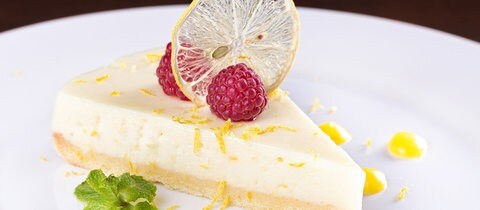 Rezept Zitronentarte mit Himbeeren: Ein Stück Zitronentarte liegt auf einem weißen Teller, garniert mit Minzeblättern