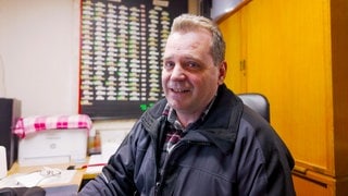Hausmeister Willi sitzt in seinem Büro im Hochhaus und lächelt in die Kamera.