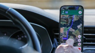 Mann tippt im Auto aufs Handy. Auf dem Handy ist eine Route von Google Maps.