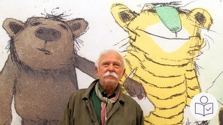 Der Künstler Janosch vor einer Wand. Auf der Wand sind Zeichnungen vom kleinen Bär und kleinen Tiger. Das Bild ist mehrere Jahre alt. Auf dem Bild ist Janosch 79 Jahre alt. Jetzt ist Janosch 95 Jahre alt geworden. Rechts das SWR Logo für Leichte Sprache.