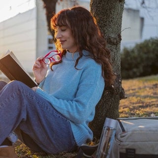 Junge Frau lehnt sitzend am Baum und liest ein Buch
