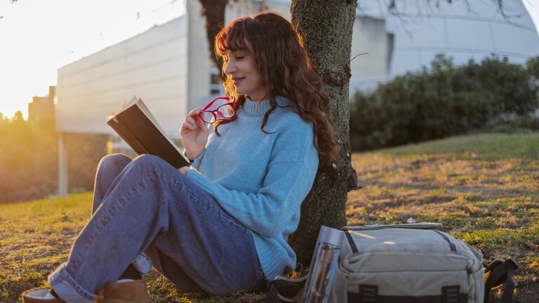 Junge Frau lehnt sitzend am Baum und liest ein Buch