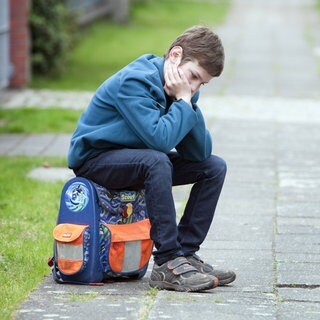 Trauriger Junge sitzt auf seinem Schulranzen: Die Schule macht Kindern und Jugendlichen zunehmend zu schaffen – eine Entwicklung, die Forschung und Medizin seit Jahren beobachten. Kopfschmerzen, Herzrasen oder Essstörungen sind nur ein Teil der Symptome. Auch Versagensangst und Mobbing in der Klasse machen Körper und Psyche krank. 
