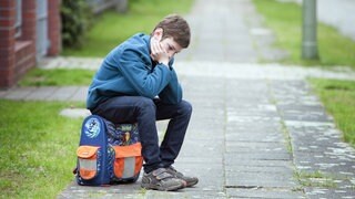 Trauriger Junge sitzt auf seinem Schulranzen: Die Schule macht Kindern und Jugendlichen zunehmend zu schaffen – eine Entwicklung, die Forschung und Medizin seit Jahren beobachten. Kopfschmerzen, Herzrasen oder Essstörungen sind nur ein Teil der Symptome. Auch Versagensangst und Mobbing in der Klasse machen Körper und Psyche krank. 