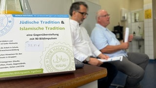 Zwei Menschen sitzen in einem Klassenraum, davor steht eine Bildersammlung mit jüdischen und muslimischen Motiven.