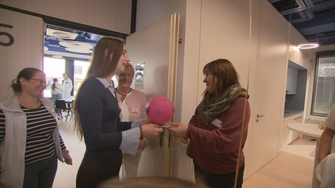 Vier Frauen mit Brille stehen an einer Tür. Eine Frau übergibt einen rosa Gegenstand. 