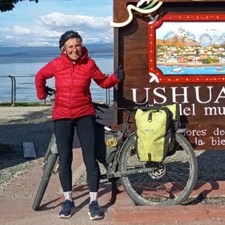 Birgit Zimmermann steht vor einem Schild in Ushuaia. Sie trägt eine rote Jacke und hält ihr Fahrrad mit einer Hand fest.
