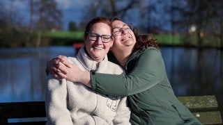 Anky (links) und Conny (rechts) sind beste Freundinnen und leben beide mit MS. Sie sind draußen und Conny umarmt Anky. Anky lacht in die Kamera. 